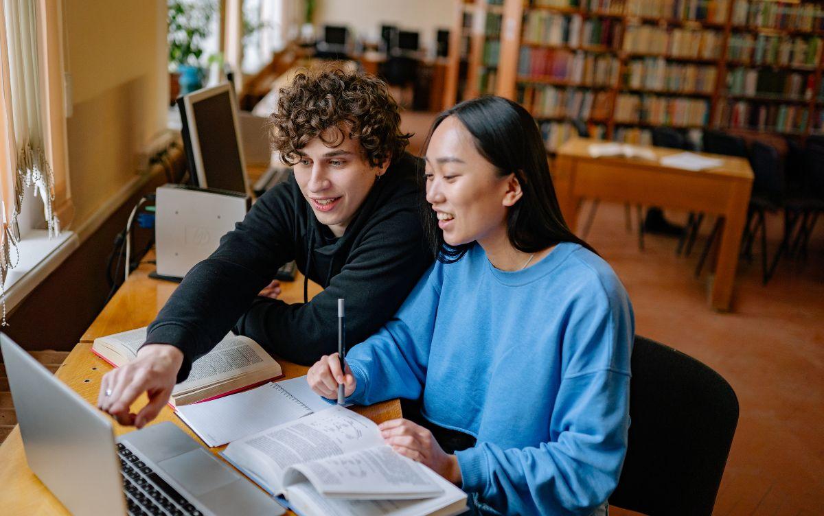Due studenti seduti in biblioteca studiano insieme consultando libri e un laptop, prendendo appunti e collaborando durante una sessione di studio condivisa