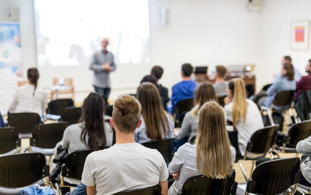 Studenti seduti in aula durante una presentazione con relatore sfocato davanti a uno schermo.